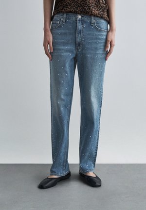 Niebieskie proste spodnie dżinsowe z małymi białymi groszkami, noszone z czarnymi balerinami i bluzką w panterkę, pokazane od talii do stóp.