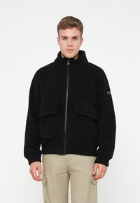 Ung man i svart fleecejacka med framfickor och beiga cargobyxor, stående mot en enkel vit bakgrund.