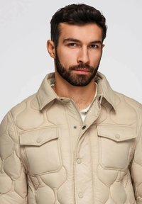 Uomo con capelli scuri e barba che indossa una giacca trapuntata beige con tasche sul petto a pattina, che guarda direttamente verso la fotocamera su uno sfondo semplice.
