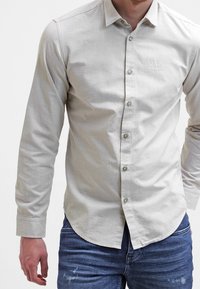 Light beige button-up shirt with long sleeves, slim fit, and subtle texture. Features a small logo on the chest and green buttons.