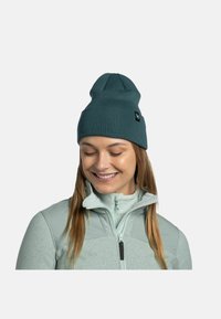 Gorro de punto en color verde azulado con la parte superior redondeada y textura de canalé. Presenta una pequeña etiqueta de marca en el lado. Se lleva con una chaqueta ligera.