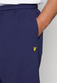 Mørkeblå svedbukser lavet af blødt stof, med en elastisk talje og et lille gult broderet logo nær lommen.