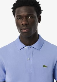 Vaaleansininen polo-paita, jossa on teksturoitu kangas, kaulus ja kaksi nappia. Paidassa on pieni vihreä brodeerattu krokotiililogo.