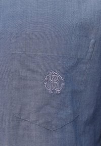 Camisa de lino azul claro con un bolsillo en el pecho. Presenta un logo bordado sutil en color a juego en el bolsillo. Acabado de tela con textura.