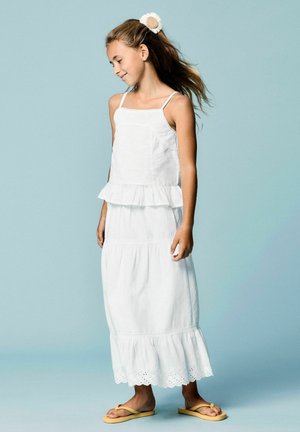 Fille en robe blanche sans manches avec des volants, épingle à cheveux fleurie et tongs jaunes, debout devant un fond bleu clair.