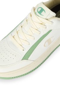 Zapatilla blanca de cuero con acentos verdes, puntera perforada, cordones planos color crema y logo de Champion. Suela de goma con agarre texturizado.