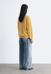 Femme aux longs cheveux noirs vue de dos, portant un pull jaune ample, un jean bleu à jambes larges et des chaussures noires pointues.