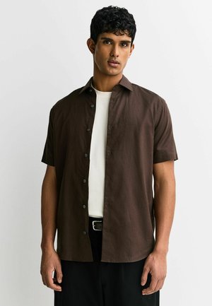 Young man with short curly hair wearing an open dark brown short-sleeve button-up shirt over a white t-shirt against a plain background.
