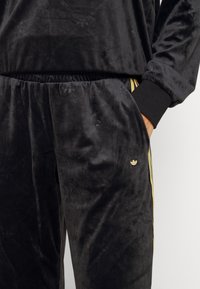 Schwarze Samt-Trainingshosen mit einem Diamantmuster, elastischem Bund, seitlichen Taschen und einem goldenen Logo-Accent an der Hüfte.