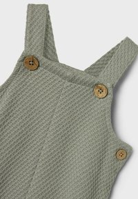 Pantalones de tirantes verdes con textura, que presentan botones de madera en los laterales y un patrón tejido a lo largo de la tela.