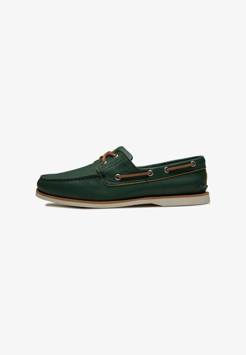 Chaussure bateau en cuir vert avec des lacets marron clair passés à travers des œillets métalliques, semelle beige et couture de style mocassin.