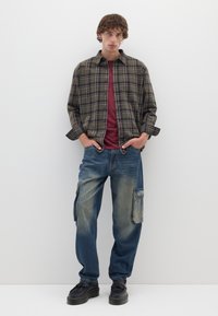 Camicia di flanella a quadri in verde scuro e beige sopra una maglietta bordeaux, abbinata a jeans cargo azzurri chiari e scarpe platform nere.