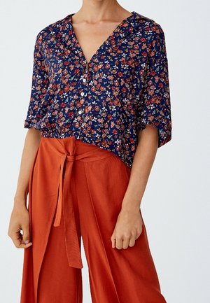 Mujer con camisa floral azul marino abotonada con mangas cortas y pantalones anchos de pierna en color naranja óxido de talle alto atados a la cintura.