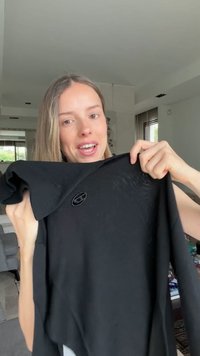 Γυναίκα μέσα σε εσωτερικό χώρο κρατάει ένα μαύρο μακρυμάνικο μπλουζάκι με μικρό κυκλικό λογότυπο κοντά στο στήθος, χαμογελάει και κοιτάζει την κάμερα.