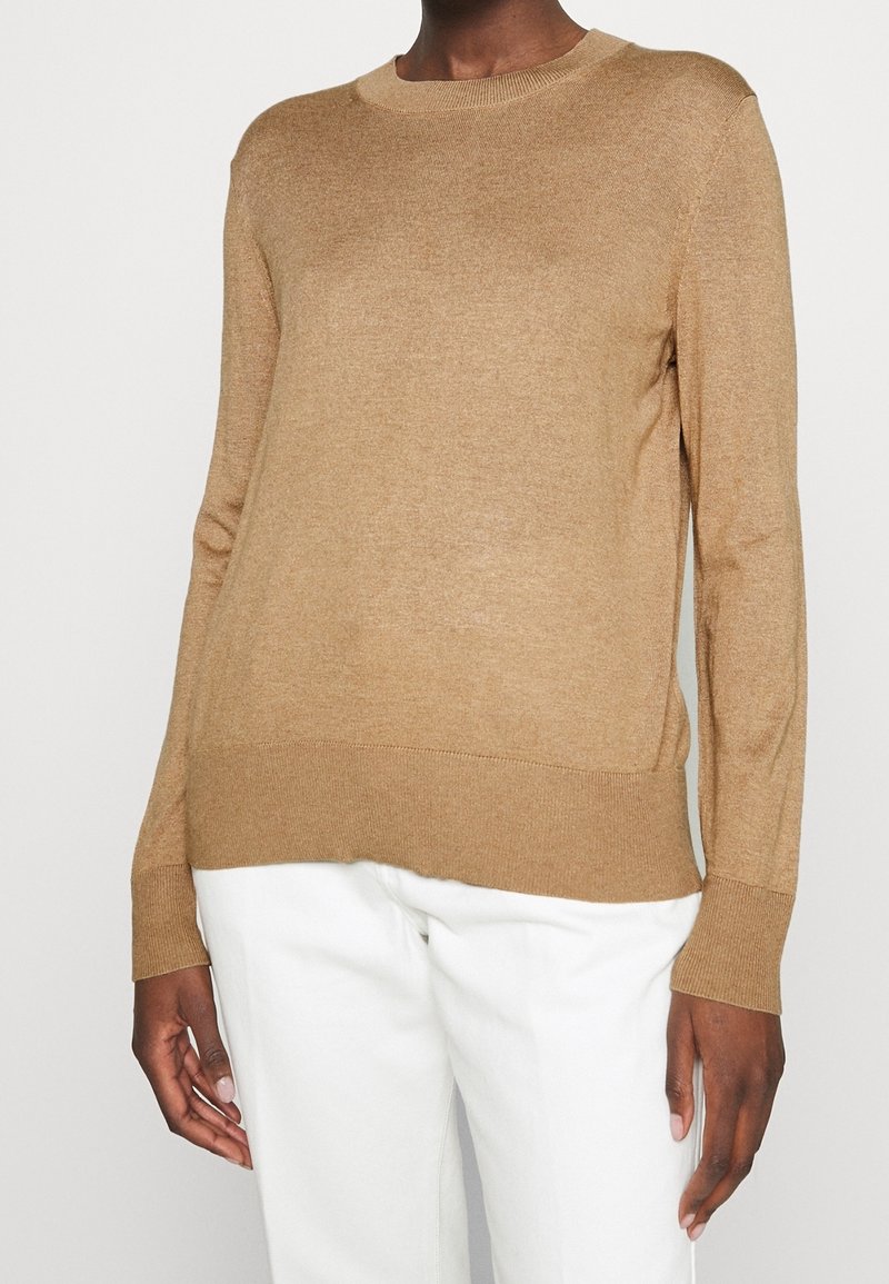 Personne portant un pull à manches longues uni couleur camel avec poignets et bas côtelés, associé à un pantalon blanc, sur un fond uni.
