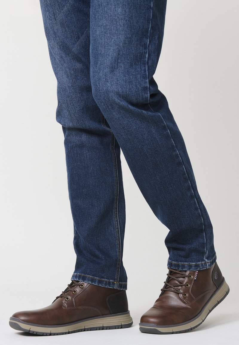 Hnedé kožené členkové čižmy s šedou gumovou podrážkou, so šnurovacím dizajnom a kontrastnými bočnými akcentmi, v kombinácii s tmavomodrými priliehavými džínsami.