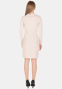 Abito aderente a maniche lunghe in tessuto beige chiaro, lungo fino al ginocchio, con scollo rotondo e piccolo dettaglio sul retro. Indossato con tacchi neri.