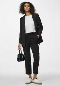 Schwarzer maßgeschneiderter Blazer mit passender Hose, weißes-cropped Hemd, hält eine kleine schwarze Handtasche und trägt weiße Sneakers mit beigen Sohlen.