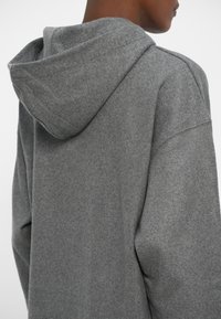 Hallika kapuutsiga dressipluus pehme ja tekstureeritud kangaga. Omab vaba lõiget ja silmapaistvat tikandit õmbluste ja kapuutsi ääres.