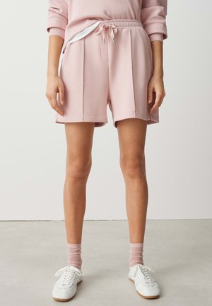 Femme portant un short rose clair à cordon, des chaussettes assorties et des baskets blanches, debout sur un sol gris contre un mur uni.