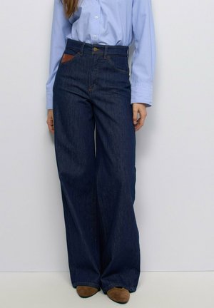 Persona con pantalones vaqueros azul oscuro de pierna ancha, camisa azul claro abotonada y zapatos marrones, de pie frente a un fondo blanco.