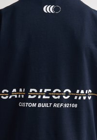 Chemise en coton bleu marine avec texte blanc et orange indiquant "SAN DIEGO INC" et "PETROL INDUSTRIES", avec un logo circulaire en haut à gauche.