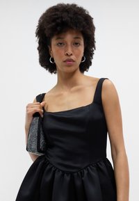 Melnas satīna kleita ar kvadrātveida dekoltē, pieguļošu augšdaļu un krokotu svārku. Modelis tur rokassomu ar pērlēm un sudraba akcentiem.