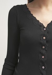 Czarny prążkowany top z długim rękawem, z ozdobnym koronkowym dekoltem w falbankę, zaokrąglonymi guzikami z przodu oraz dopasowaną fakturą.