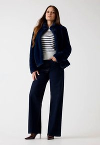 Navy fur jacket with a high collar, paired with a striped sweater and wide-leg dark jeans. The model wears pointed-toe heels.