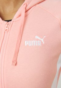 Svetlo roza zip-up kapuca iz mehkega materiala, z kenguru žepom in belim vezenim Puma logotipom na prsih.