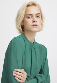 Blusa verde con un collo alto e dettagli arricciati, realizzata in tessuto leggero, con maniche lunghe e polsini elasticizzati.