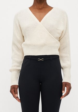 Person wearing a cream-colored knitted wrap sweater with long sleeves and black high-waisted pants with a slim black belt.