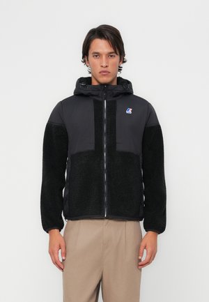 Veste noire avec un design matelassé et une texture duveteuse sur les manches et le bas du corps. Comprend une capuche et une fermeture éclair devant.