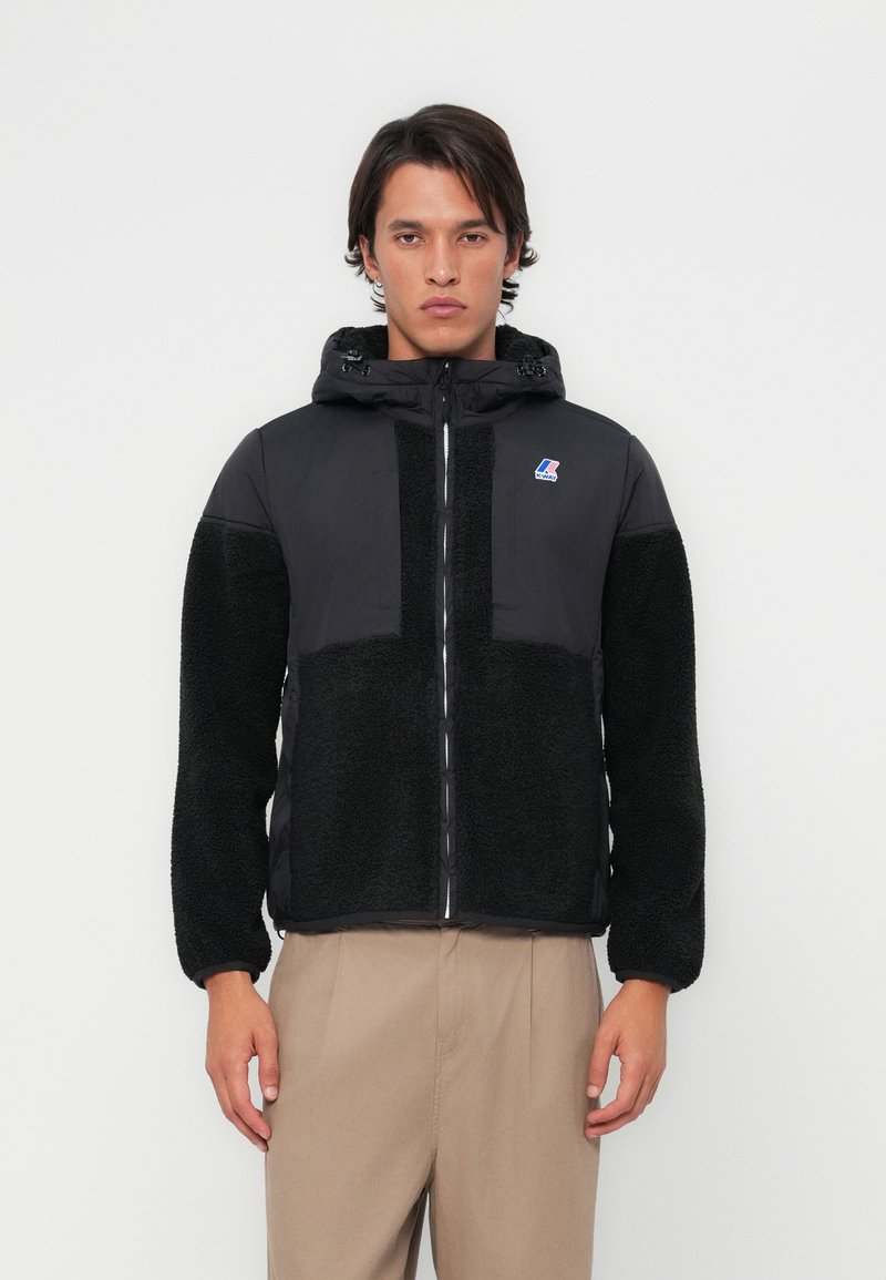 Veste noire avec un design matelassé et une texture duveteuse sur les manches et le bas du corps. Comprend une capuche et une fermeture éclair devant.