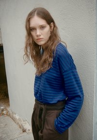 Jonge vrouw met lang golvend haar, gekleed in een blauw gestreepte trui en bruine broek, staand met haar handen in haar zakken tegen een gestructureerde muur.