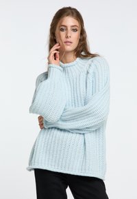 Light blue oversized knitted sweater with ribbed texture, wide sleeves, and a round neckline. Paired with dark pants for contrast.