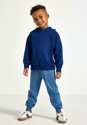 Laivastonsininen hupullinen collegepaita, siniset jogger-housut ja valkoiset lenkkarit mustilla yksityiskohdilla. Pehmeä kangas, rento istuvuus, resorit hihansuissa ja helmassa.