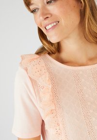 Top rosa claro con detalle de encaje con volantes en el hombro, con patrones texturizados y recortes florales intrincados en toda la tela.