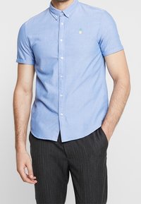 Camicia a maniche corte azzurra chiara con bottoni, realizzata in un misto cotone, con colletto. Presenta un piccolo ricamo di un ananas verde sul lato sinistro del petto.