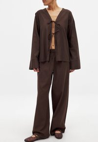 Rjava dvodelna obleka, ki vključuje bluzo z dolgimi rokavi in V-izrezom ter zaveznico, v kombinaciji s širokimi hlačami. Gladko, lahko blago.
