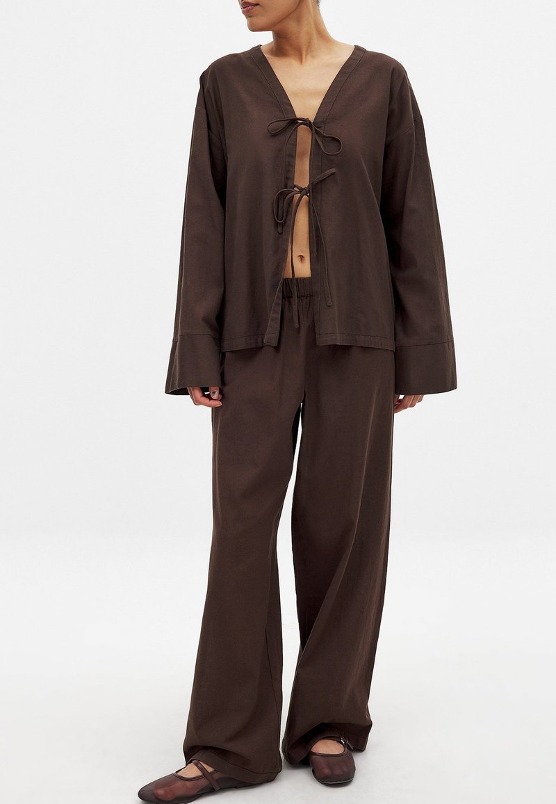 Rjava dvodelna obleka, ki vključuje bluzo z dolgimi rokavi in V-izrezom ter zaveznico, v kombinaciji s širokimi hlačami. Gladko, lahko blago.