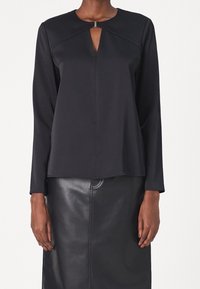 Haut noir à manches longues avec une ouverture en trou de serrure à l'avant, tissu lisse et encolure structurée agrémentée d'un détail métallique, associé à une jupe en cuir élégante.