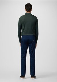 Top de manga larga verde oscuro con cuello alto, combinado con pantalones azul marino que tienen un bolsillo trasero y un detalle de logo. Adecuado para uso casual.