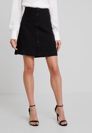 Kvinde iført sort knap-front nederdel, hvid langærmet bluse og sorte høje hæle sandaler mod en ensfarvet baggrund.