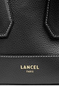 Sac à main en cuir noir avec une texture granuleuse, présentant des coutures blanches et un logo doré en relief portant la mention "LANCEL PARIS."