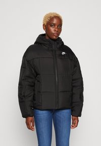 La veste matelassée noire présente une texture quiltée, une capuche ajustable, une fermeture éclair à l'avant et le logo Nike sur la poitrine gauche. Associée à un jean bleu.