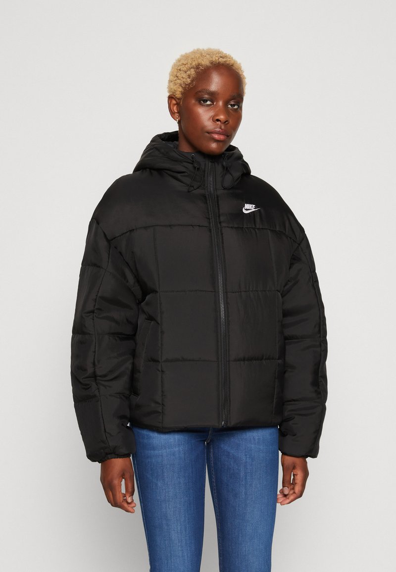 La veste matelassée noire présente une texture quiltée, une capuche ajustable, une fermeture éclair à l'avant et le logo Nike sur la poitrine gauche. Associée à un jean bleu.