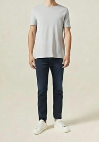 Homme portant un t-shirt à manches courtes gris clair, un jean slim foncé et des baskets blanches, debout devant un fond beige uni.