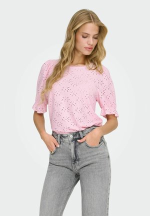 Kobieta o długich blond włosach, ubrana w różową bluzkę z ażurowym wzorem z krótkim rękawem oraz wysokim stanem szare dżinsy, z rękami w kieszeniach.