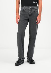 Jeans de denim negro con diseño de pierna recta, que presentan un acabado desgastado, cinco bolsillos y un cierre de botón metálico. Llevados con mocasines negros.